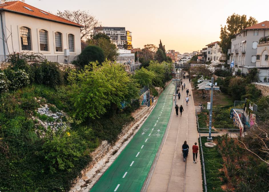 אנשים הולכים ורוכבים על אופניים לאורך שביל עירוני ירוק המוקף בעצים ובניינים בשעת השקיעה.