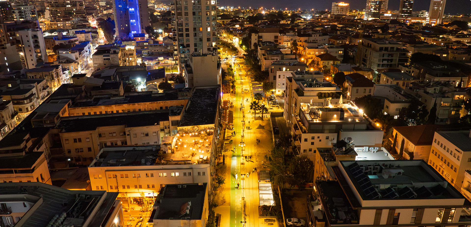 מבט לילי אווירי על רחוב עירוני מואר באור בהיר, משובץ בבניינים ובאנשים הולכים.