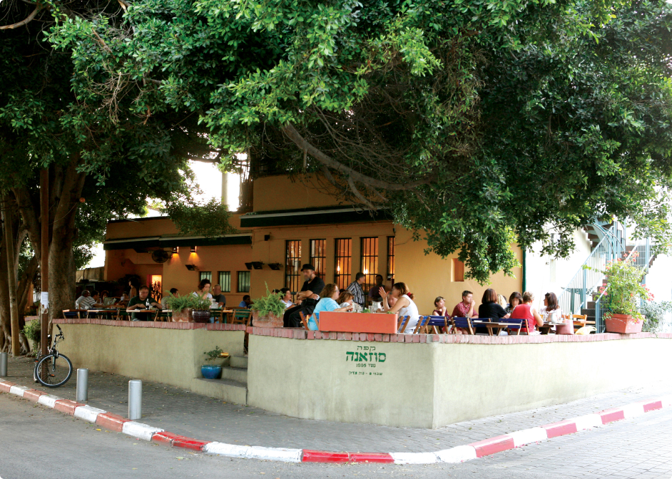 אנשים סועדים בשולחנות בחוץ תחת עצים גדולים ליד בניין צהוב בפינת רחוב בעיר.