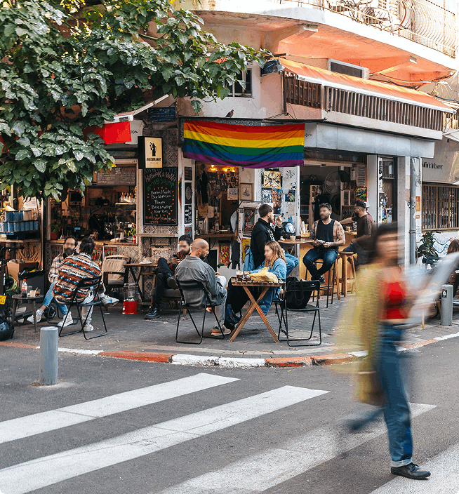 אנשים יושבים מחוץ לבית קפה הומה אדם, שמעליו מתנוסס דגל הקשת, בעוד אדם חוצה את הרחוב מולו.