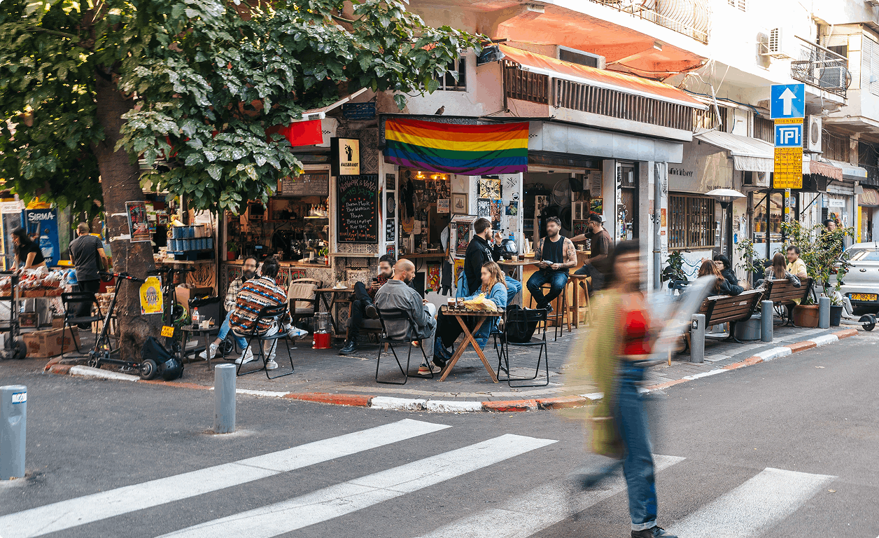 אנשים יושבים בשולחנות בחוץ ליד בית קפה עם דגל הקשת; אדם חוצה את הכביש במעבר חצייה שנמצא מול בית הקפה.
