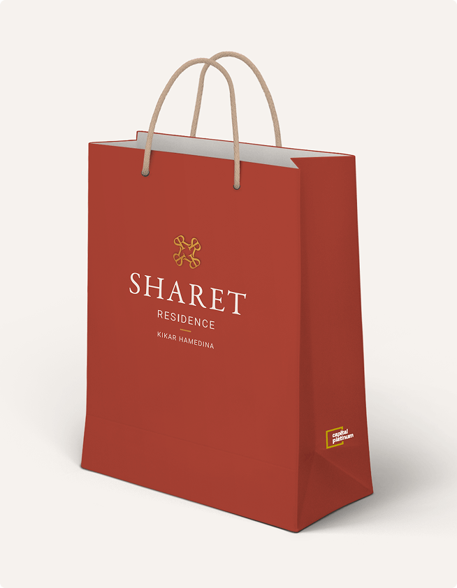 שקית קניות אדומה עם הלוגו "SHARET RESIDENCE", ידיות בצבע בז' ותווית סגולה קטנה בתחתית.
